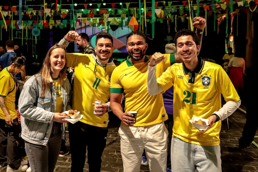 Three men and one woman wearing bright yellow and green Brazil outfits inside a dark event room full of people and decorations
