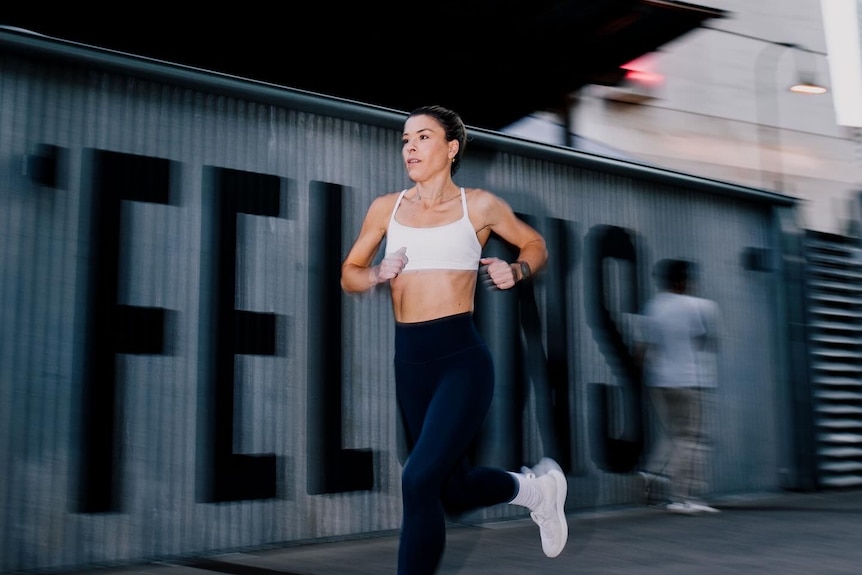 A dark-haired woman in a sports bra and leggings runs down a city street.