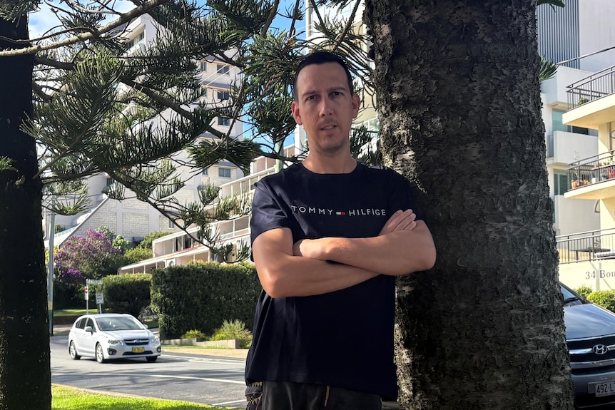 Leigh Peters leans against a tree with his arms crossed.