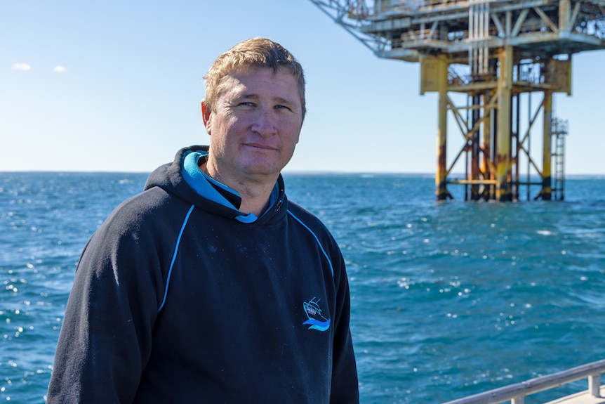 Mike is standing on a boat with the platform behind him. He is wearing a navy blue jumper.