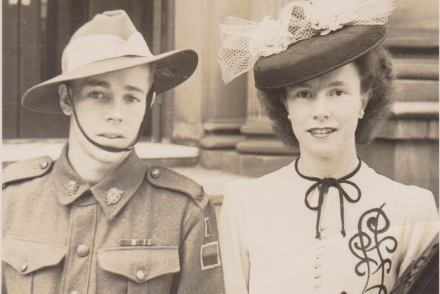 A soldier in world war two uniform stands next to a woman wearing a hat with a veil and flowers on it.