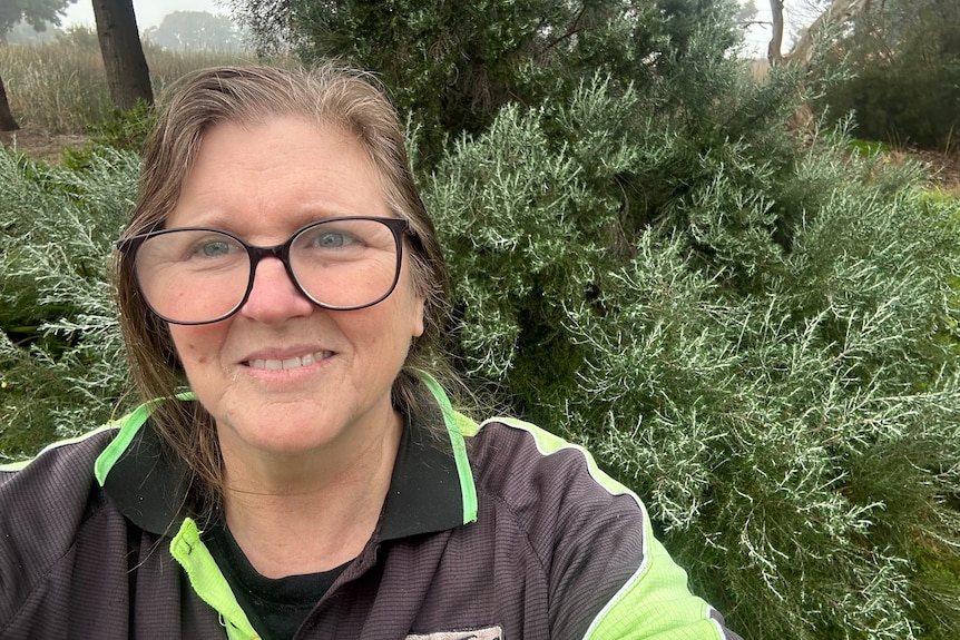 A woman wearing glasses smiling at the camera with a green bush in the background