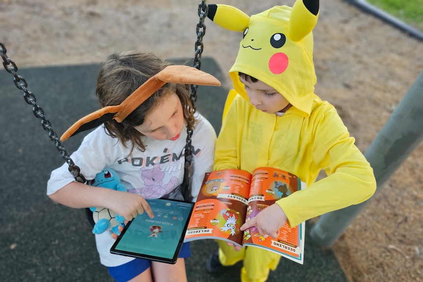 Two kids in pokemon gear on a swing set consulting a pokemon book