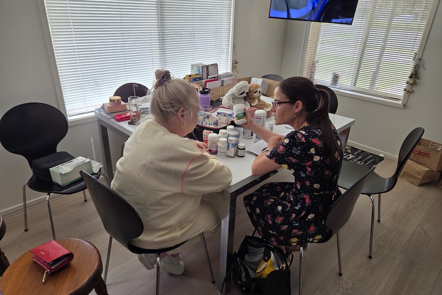 Two women sit at a table full of medications and talk. They are in a house.