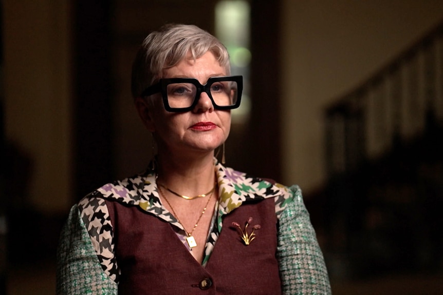 A woman wearing black-rimmed spectacles is interviewed for television.