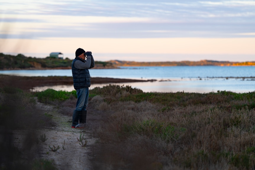 A man with a beanie on stands on the banks of the Coorong and raising binoculars to look out at the water