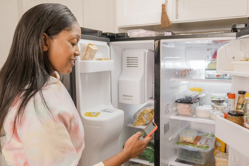 A woman stands in front of open freezer and fridge full of food, looking at her phone.