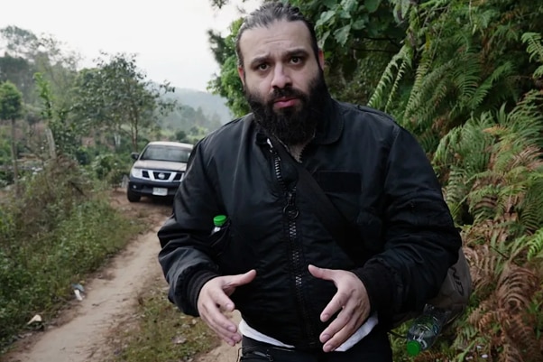 A man with a beard and black jacket standing in tropical bushland.