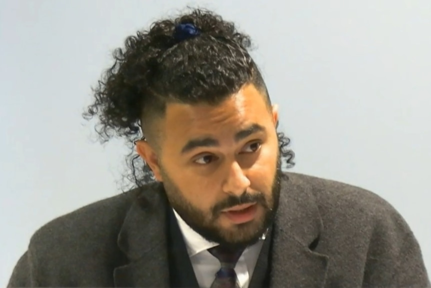 A man with tied up curly hair and a beard in a grey coat, giving evidence.