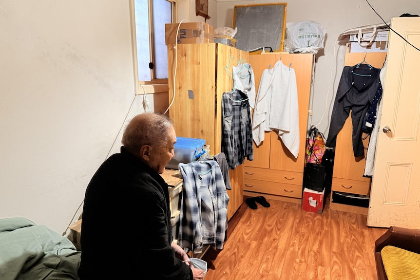 A man sitting on a bed with his back to the camera with cupboards against the far wall with clothes hanging off them