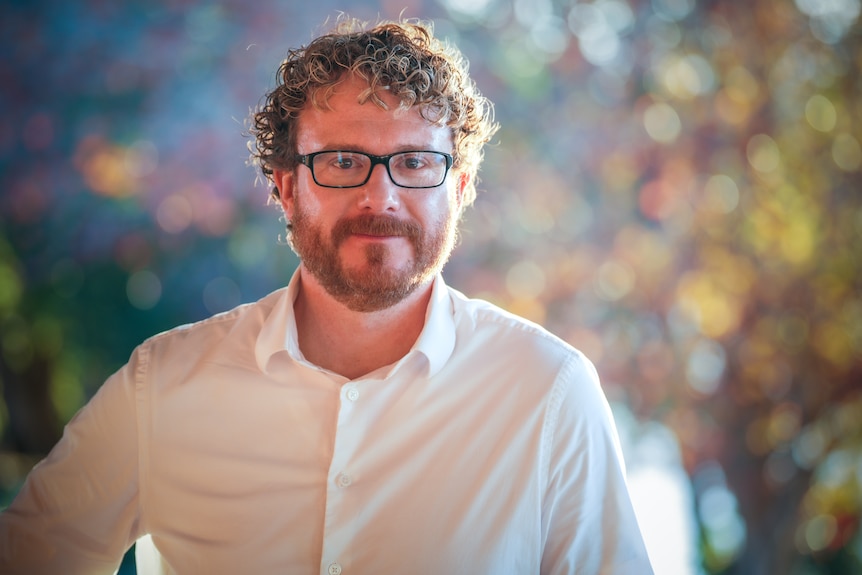 A man with curly hair, black-framed glasses wearing a white business shirt.