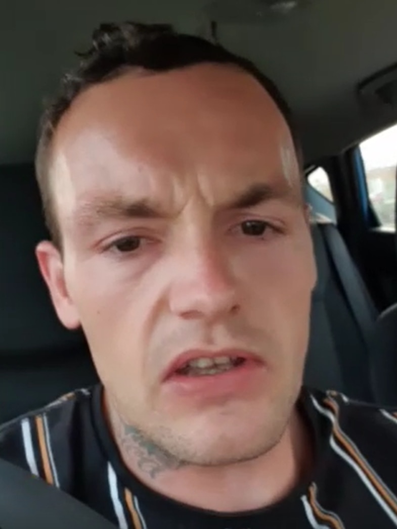 A dark haired man in a black, white and beige striped shirt stares at the camera.