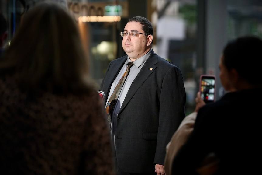 Andre Oboler in a suit, tie, glasses, speaking to a crowd outside an office building.