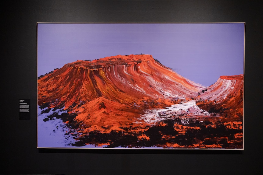 A large painting hangs on a wall inside an art gallery, it depicts a rocky landscape with orange and purple hues.