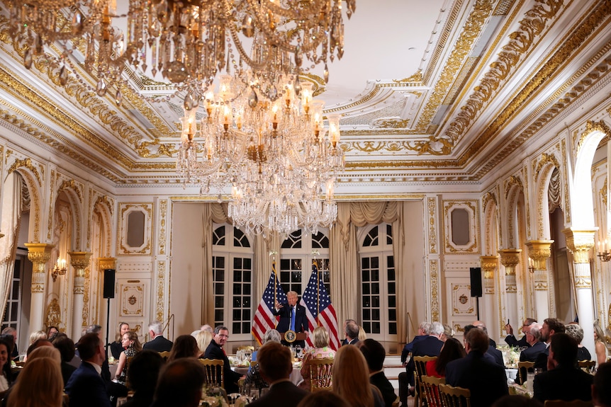 A wide shot of Donald Trump speaking in a grand ballroom decorated with gold with a crystal chandelier