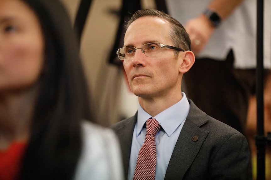 A man in a suit and glasses looks ahead while sitting near other people.