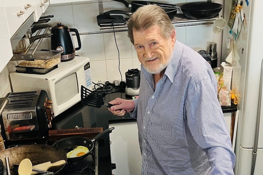 A man smiles while posing for a photo in his kitchen frying an egg.