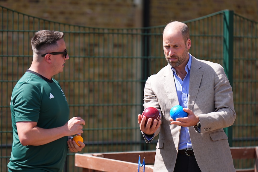 Prince William holds two bowls as another man shows him what to do.