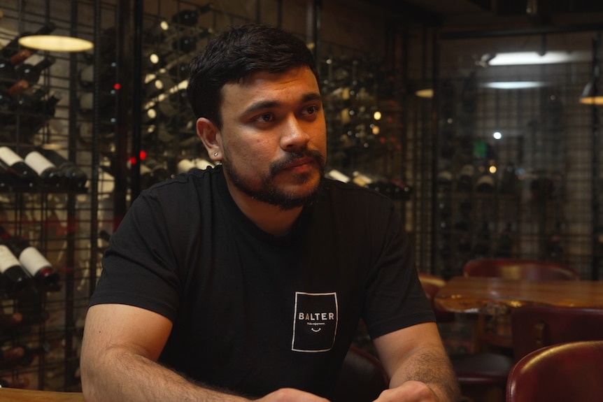 Dinesh Khadka wears a black t-shirt and sits at a table in front of metal shelving containing bottles of wine.
