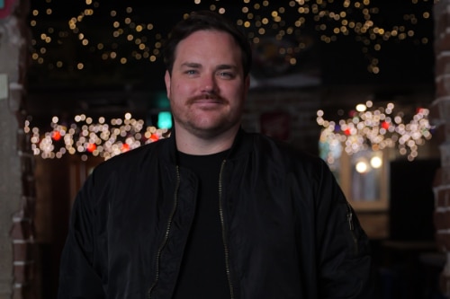 A young man in a dark jacket inside a room decorated with fairy lights.