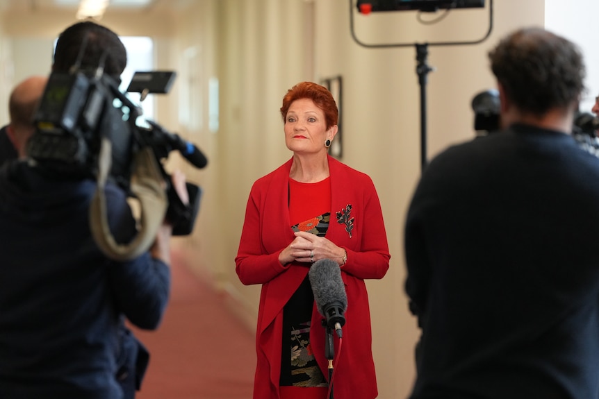 A woman in a red, with red hair, standing in a corridor in front of TV cameras.