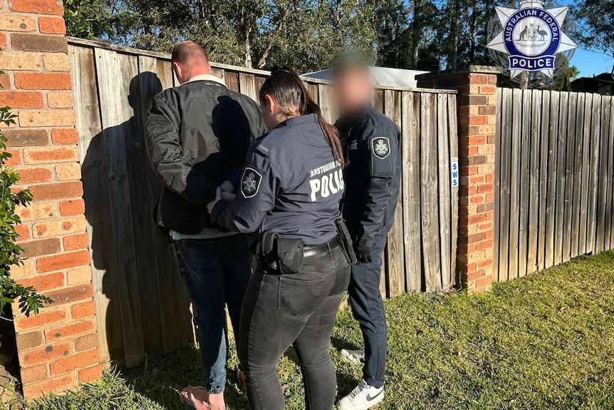 former childcare worker up agianst a wooden fence as he is being handfuffed by two australian federal police officers