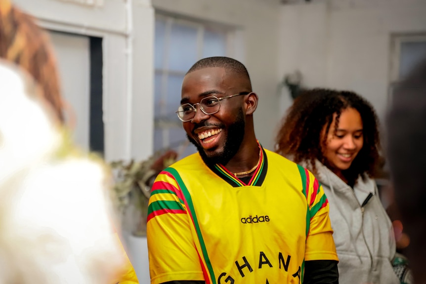 A man wearing a yellow soccer jersey with red and green stripes on the sleeves smiles with his mouth open. He is wearing glasses