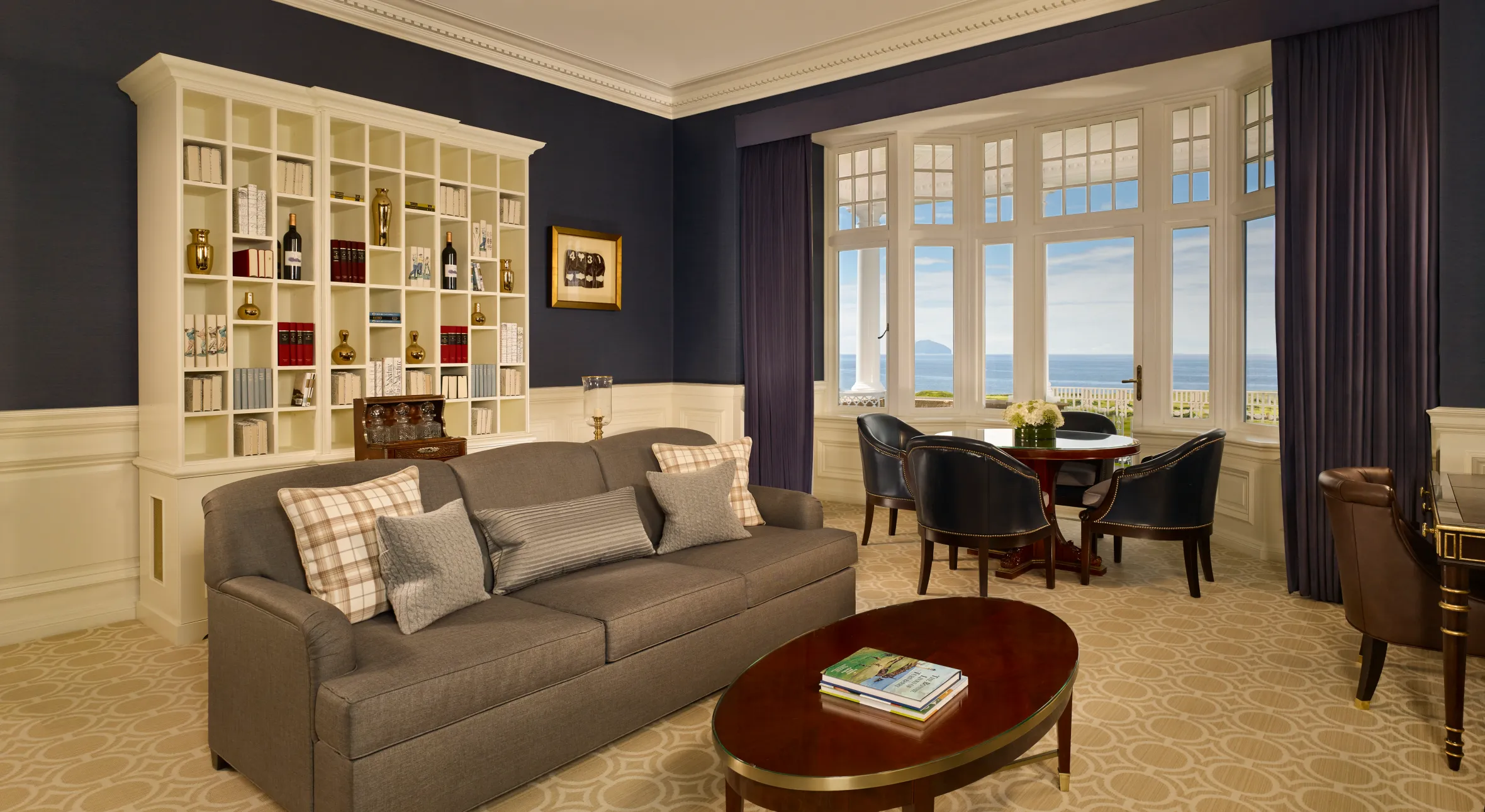 A luxury room at Trump Turnberry with a dark blue wall, a bookshelf, a grey sofa, and a bay window overlooking the ocean.