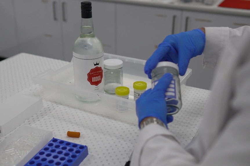 A person opening up a testing jar with a large bottle of alcohol sitting in a plastic tray on a table.