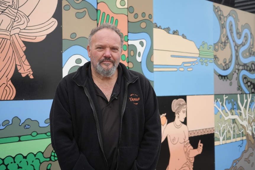 A grey-haired bearded man, in black with Venus Citrus logo in front of a mural featuring the river murray and Greek statues.
