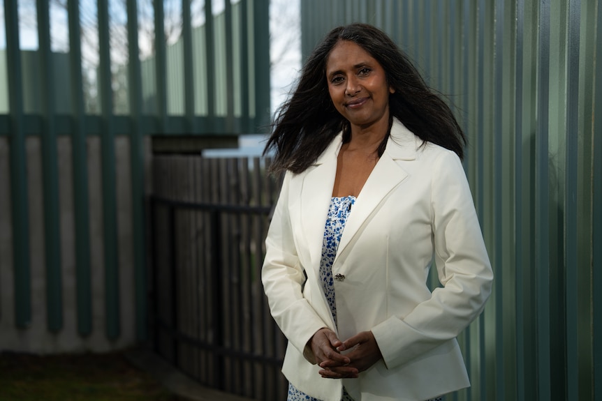 A woman with black hair wears a light grey winter coat and stands in front of a microphone in a park