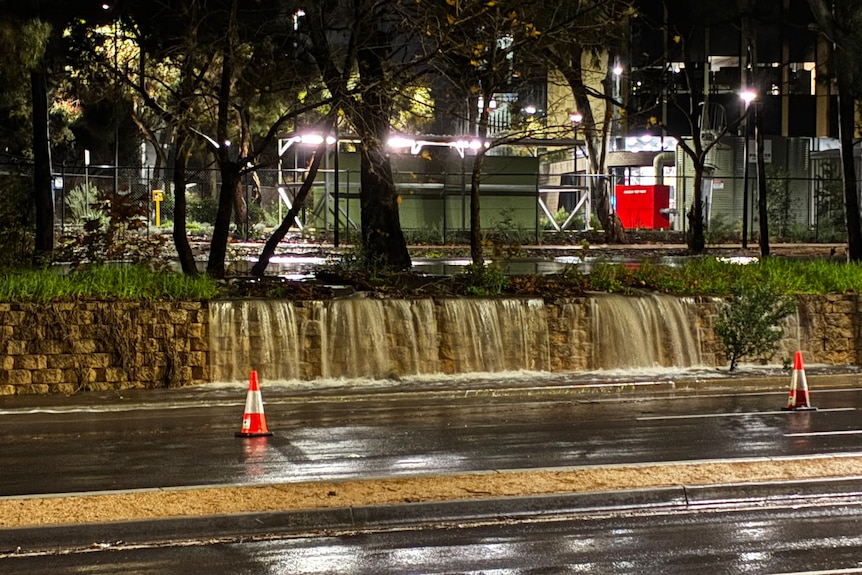 water overflow a short wall next to a road partially blocked by traffic cones