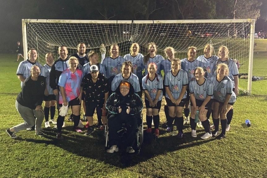 A soccer team standing in two rows with a soccer goal behind. A boy is in front, in a wheelchair.