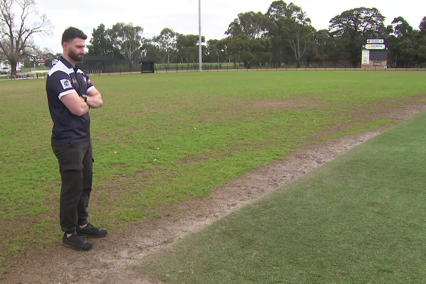 A dark haired man with a beard wearing a dark top and pants looks down at artificial turf in a footy oval.