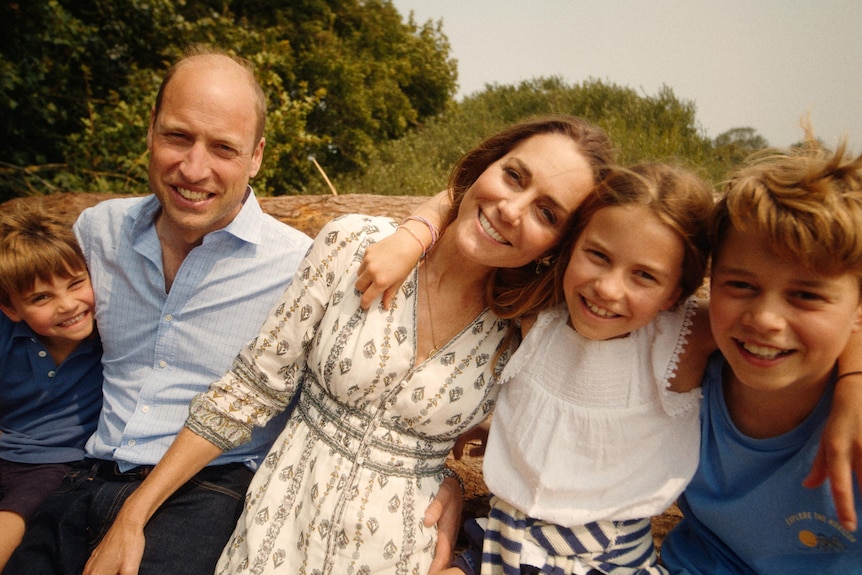 Two boys, a girl, a man and a woman sit together with arms around each other smiling with green trees, countryside behind them