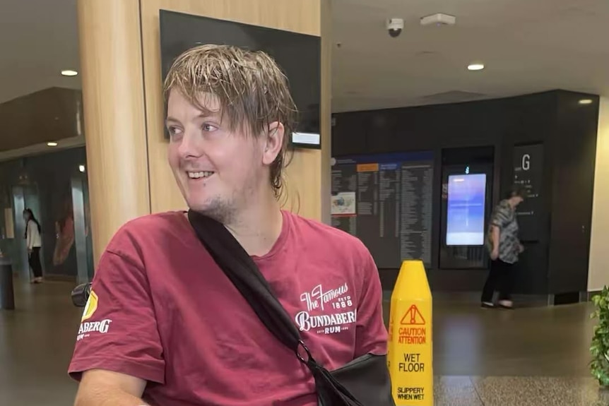 A smiling young man with dark hair and his arm in a sling sits in a mall.
