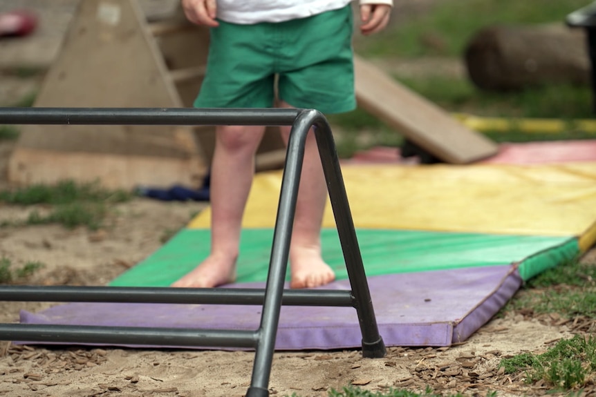 A child stands on a coloured mat in front of a small piece of climbing equipment. They are seen from the waist down.