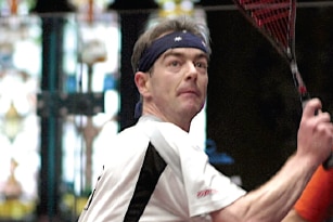 A middle aged man in white prepares to play squash backhand on a court