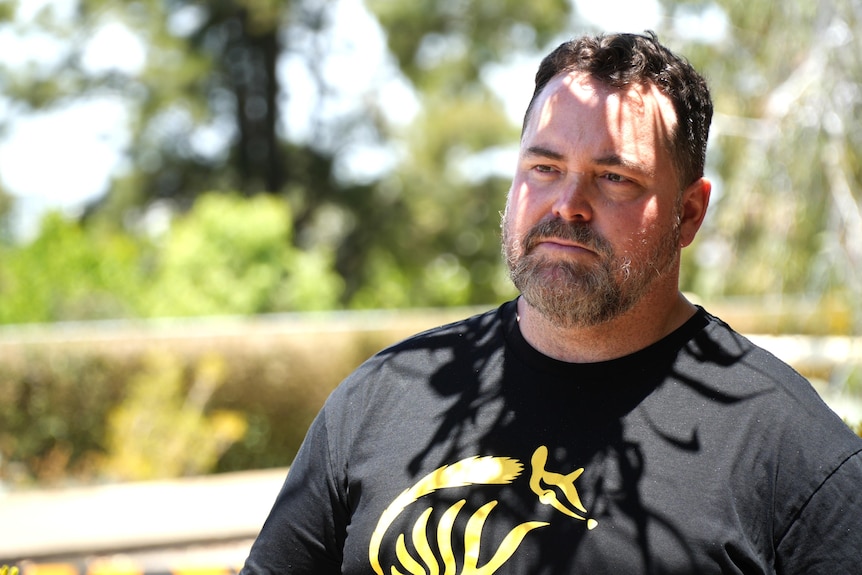 Matt Roberts stands outdoors wearing a black t-shirt with a yellow logo on it.