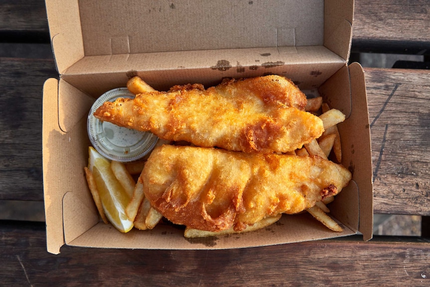A cardboard takeaway box on a picnic table holds golden, fried fish atop chips with a slice of lemon and tartare sauce.