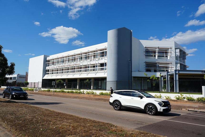 A three storey white building viewed from across a road with a white sedan driving past