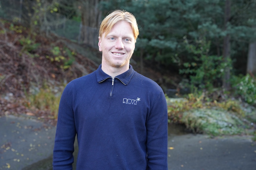 A young man wearing an NRM South blue quarter-zip jumper stands beside a stream at Hobart Rivulet.