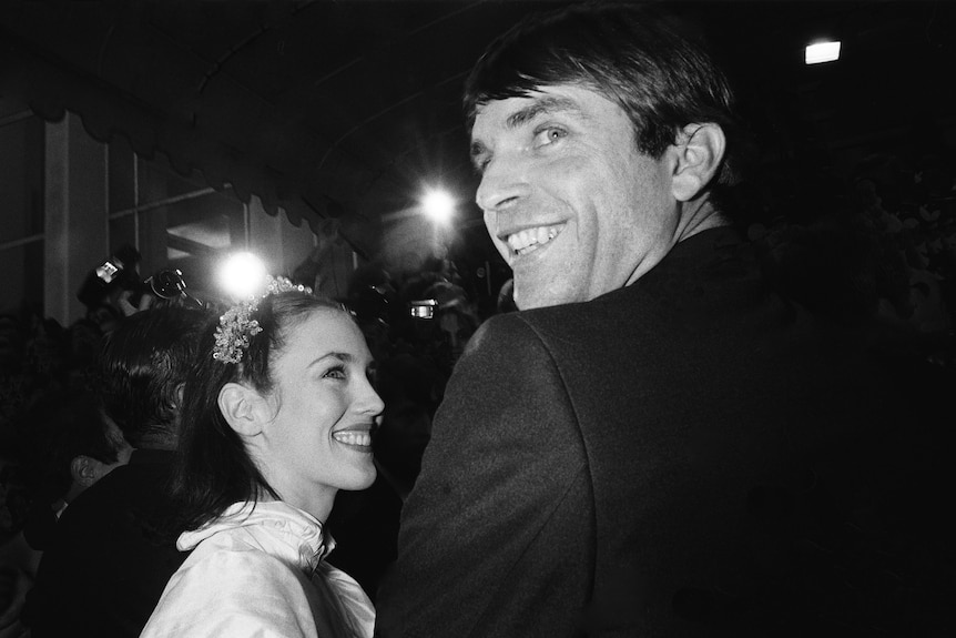 black and white photograph of Sam Neill in 1981 turning back and looking at cameras on the red carpet. A female is next to him