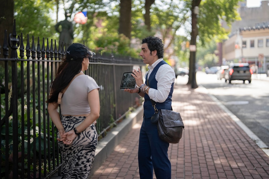 A man with curly hair chats to a woman while holding up a photo of Taylor Swift on an Ipad.