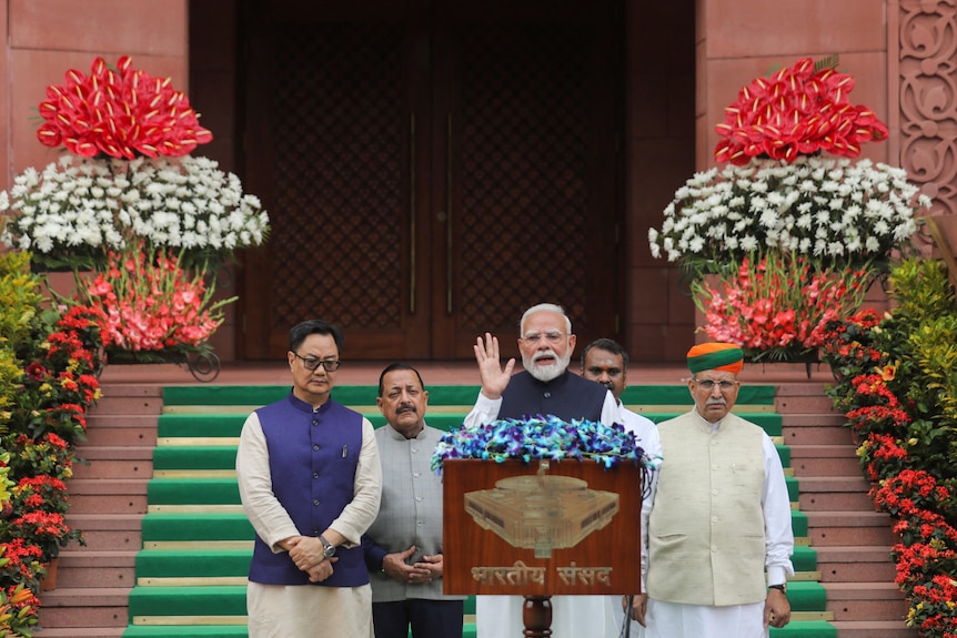 Narendra Modi lifts up one hand as he speaks at a podium in front of parliament steps, four men standing behind him