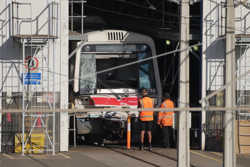 A damaged Transperth passenger train carriage parked in a shed of a depot with workers in hi-vis vests standing nearby.