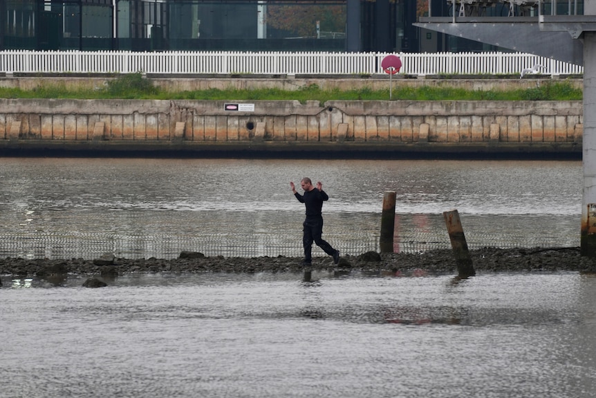 A man dressed in a black top and pants walks along the rocks protruding from a river with his hands in the air.