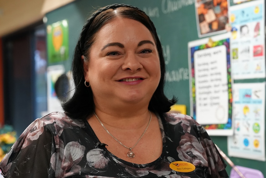 A woman stands in front of a blackboard covered in posters. She has dark shoulder length hair and a headband.