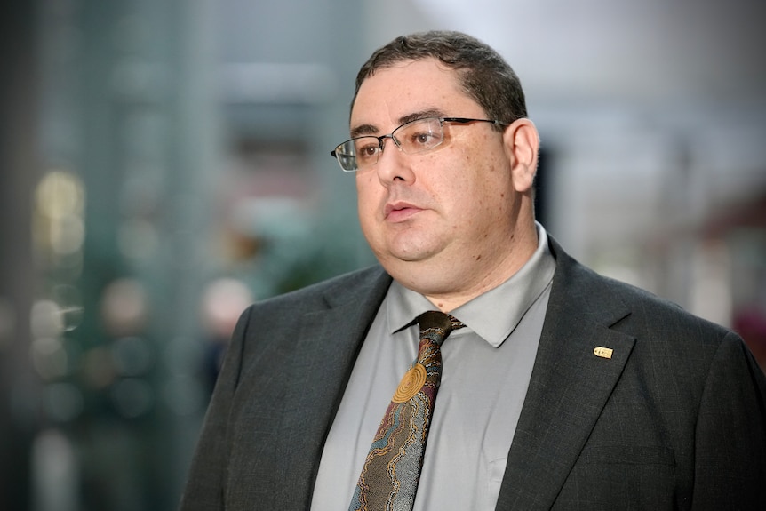 Andre Oboler in a suit, tie, glasses, speaking to a crowd outside an office building.