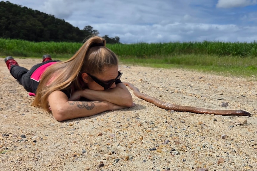 A young woman lies on the ground with a snake stretched out beside her.